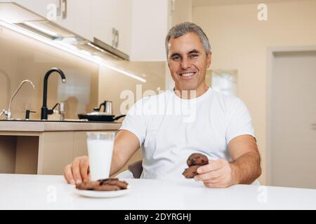 Frühstückskonzept. Fröhlicher älterer Mann in weißem T-Shirt hat ein Glas Milch und Schokoladenkekse für ein Frühstück heute. Smart Küche Interieur mit Stockfoto