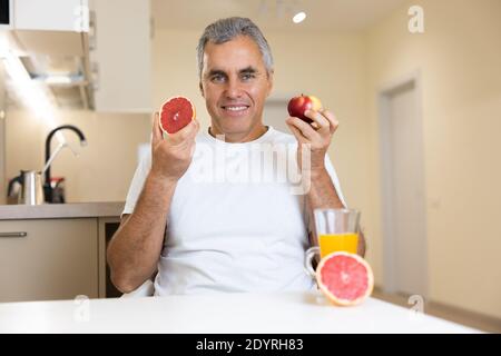 Gut aussehende reife Mann isst Früchte. Bio gesunde Ernährung Konzept. Fröhlicher Mann, der Apfel und Grapefruit in der Nähe des Gesichts hält und vor der Kamera lächelt. Glas Stockfoto
