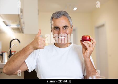 Erwachsene Mann bevorzugt gesunde Ernährung. Helles, intelligentes Interieur auf dem Hintergrund. Reifer Mann hält roten frischen Apfel, Daumen hoch und lächelt mit Zähnen. Der Mann sitzt da Stockfoto