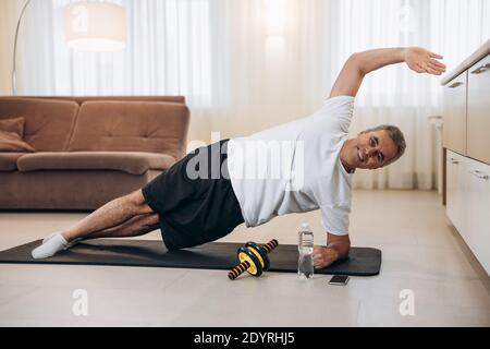 Glücklicher älterer Mann, der mit erhobener Hand eine seitliche Plankenübung macht, lächelt und die Kamera anschaut. Gesundes Lifestyle-Konzept. Achten Sie auf Ihren Körper. Alt Stockfoto