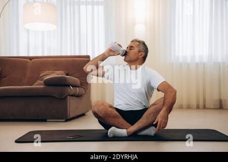 Älterer Mann trinkt nach dem Yoga Wasser und sitzt mit gekreuzten Beinen. Schwarze Yogamatte. Der Mann ist nach dem Training sehr müde. Trinken Sie mehr Wasser. Elegantes, modernes Zimmer Stockfoto