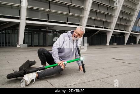 Guy fiel während der Fahrt mit dem E-Scooter schnell. Stylischer Mann in grauem Hoodie sitzt auf dem Boden und hat Knieschmerzen. Grüner Elektroroller. Umweltfreundlich Stockfoto