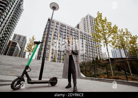 Frau hat sich einen Roller gemietet. Herbst in der Großstadt. Attraktive Frau in Freizeitkleidung diskutiert Bedingungen der Vermietung Roller von seinem Smartphone. Appartementblöcke weiter Stockfoto