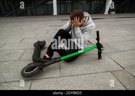 Seien Sie vorsichtig Guy fiel während der Fahrt die Elektroroller schnell. Der Mann im grauen Kapuzenpulli sitzt auf dem Boden und hat Kopfschmerzen. Umweltfreundliches Transportkonzept Stockfoto