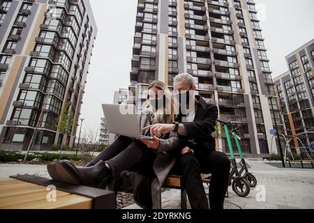Junges Paar sitzt auf Bank im Park in der Nähe ihrer Wohnung. Sie tragen schwarze medizinische Gesichtsmasken und halten Abstand zu anderen Menschen. Kümmern Sie sich um Stockfoto