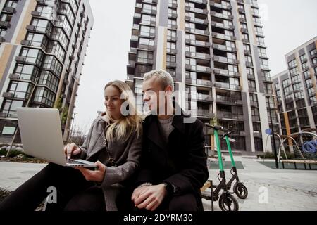Junge Frau zeigt Fotos ihrem männlichen Freund und lächelt. Blonde Mann sitzt in der Nähe und auf Laptop. Sie sitzen auf einer Bank in der Nähe von Wohnblöcken Stockfoto