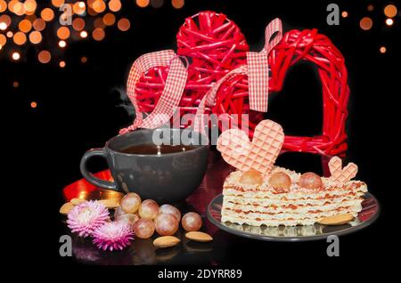 Waffelkuchen mit Herz und Trauben und heißem Tee in einer Tasse dekoriert. Schwarzer Hintergrund mit Bokeh und Herzen. Valentinstag-Konzept, selektiver Fokus Stockfoto