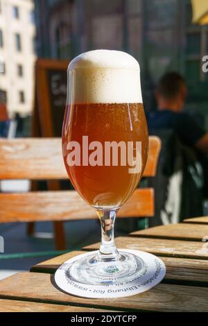 Ein Glas frisches, braunes Bier mit Schaum darauf Auf Holztisch mit Gebäuden der Stadt im Hintergrund Stockfoto