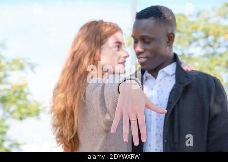 Afrikanischer schwarzer Mann und rothaarige kaukasische Frau, die einen Verlobungsring an die Kamera zeigt und sich gegenseitig anschaut. Heiratsantrag. Stockfoto