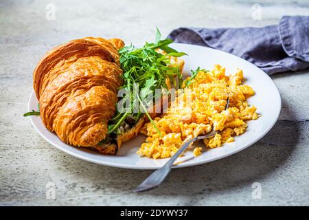 Rührei und Croissant mit Käse und Rucola auf einem weißen Teller. Eier und Croissants zum Frühstück. Frühstückskonzept. Stockfoto