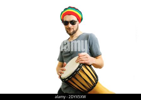Junger kaukasischer lächelnder Mann in Rasta-Hut, Sonnenbrille und grauem T-Shirt auf weißem Hintergrund mit Djembe afrikanischer Trommel. Stockfoto