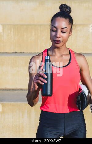 Vertikales Porträt eines attraktiven afro-amerikanischen Athleten, der Boxen hält Handschuhe und eine schwarze Metallflasche für Wasser Stockfoto