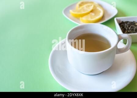 Weiße Tasse mit grünem Tee auf der Oberseite eines Grüner Tisch mit einem Teller mit Zitronenscheiben und s Kleine Untertasse mit trockenen grünen Teeblättern auf dem Hintergrund Stockfoto