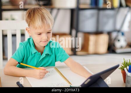 Smart Schuljunge beobachten Lektion online auf Tablet-Computer und nehmen Notizen im Notizbuch beim Lernen von zu Hause Stockfoto