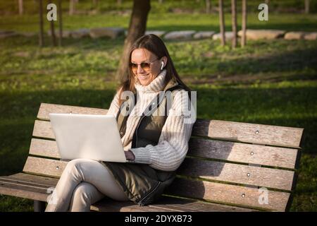 Glückliche Frau, die im Stadtpark mit Laptop und Kopfhörern arbeitet. Student, der im Freien studiert. Freiberuflich tätig Stockfoto