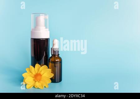 Ätherisches Öl in kleiner offener brauner Tropfflasche mit liegender Glaspipette, große Flasche mit weißem Spender und gelber Blume auf blauem Hintergrund. Konz Stockfoto