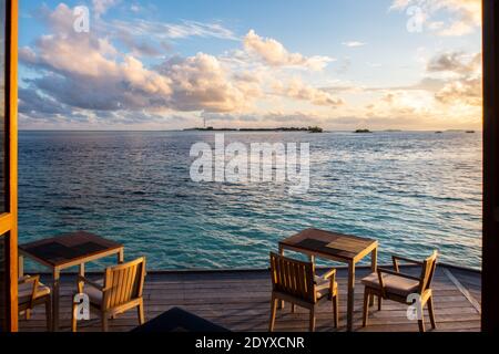 Terrasse mit hölzernen Esstischen über dem türkisfarbenen Meer, mit Wolken und Sonnenuntergang im Hintergrund, Malediven. Stockfoto