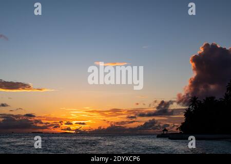 Dramatischer Sonnenuntergang über dem Indischen Ozean auf der Insel Vilamendhoo auf den Malediven, mit Cumulonimbuswolken und Silhouette eines Holzbungalows und Palmen. Stockfoto