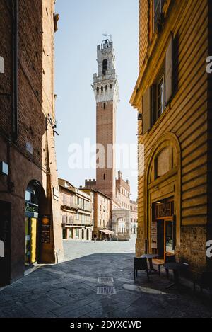 Blick von einer schattigen Gasse in der Altstadt von Siena auf den in der Sonne scheinenden Turm des Palazzo Publico auf der Piazza del Campo, Toskana, Italien Stockfoto