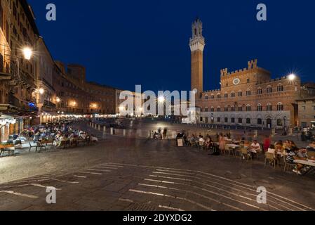 Menschen sitzen zum Abendessen in den Restaurants auf dem mittelalterlichen Platz Piazza del Campo vor dem Palazzo Pubblico in Siena, Toskana, Italien Stockfoto