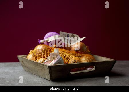 ZUTPHEN, NIEDERLANDE - Dec 16, 2020: Low-Key-Stillleben von hölzernen Container-Schale Kiste mit Zwiebeln in einem Netz und Knoblauch auf einem rustikalen Küchentheke wit Stockfoto