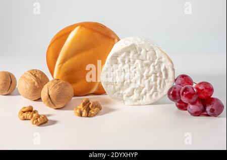 Zwei ganze Köpfe, Käserad, Brie und geräucherter Käse suluguni mit Walnuss und Traube auf weißem Hintergrund. Nahaufnahme Stockfoto