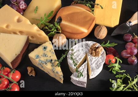 Käsehintergrund. Sortiment verschiedene Sorten Käse mit Walnüssen, Tomaten und Trauben auf schwarzem Schiefer Hintergrund. Blauschimmelkäse Dorblin, geräuchert, Brie, Maas Stockfoto