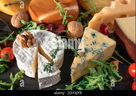 Käsehintergrund. Sortiment verschiedene Sorten Käse mit Walnüssen, Tomaten und Trauben auf schwarzem Schiefer Hintergrund. Blauschimmelkäse Dorblin, geräuchert, Brie, Maas Stockfoto