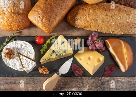 Verschiedene Käse auf schwarzem Stein Brett ausgelegt. Blauschimmelkäse Dorblin, geräucherte Suluguni, Brie, Maasdam, Cheddar, Parmesan mit Walnüssen, Trauben, Tomaten und Stockfoto