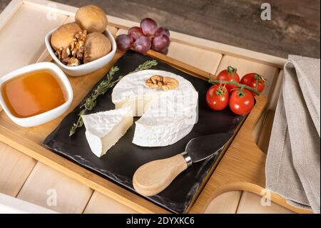 Holztablett zum Frühstück mit Brie-Käse, Honig, Walnüssen, Tomaten. Käse Frühstück auf dem Holztablett mit schwarzem Teller Stockfoto