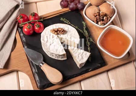 Holztablett zum Frühstück mit Brie-Käse, Honig, Walnüssen, Tomaten. Käse Frühstück auf dem Holztablett mit schwarzem Teller Stockfoto