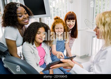 Kinder und Mütter in der Zahnarztpraxis. Zwei multiethnische Mütter mit ihren Töchtern im Teenageralter sitzen im Stuhl der Zahnmedizin und zeigen auf einen digitalen Tablet-pc Stockfoto