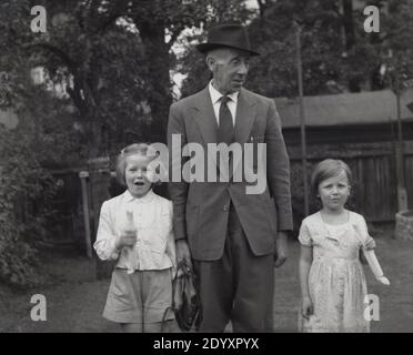 1950er Jahre, historisch, zwei junge Mädchen mit ihrem Großvater, der Anzug und Krawatte und einen dreifachen Hut trägt. Die jungen Mädchen tragen etwas, das aussieht wie Steinstöcke, ein beliebtes Süßes in dieser Zeit. Stockfoto