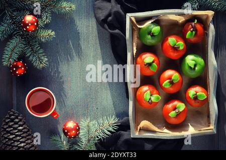 Marzipan Schwamm Äpfel. Weihnachtsdessert mit Tasse Tee, Tannenzweigen, grüner Kerze, Tannenzapfen, Weihnachtsspielzeug. Draufsicht auf rustikales Holztablett auf Textil Stockfoto