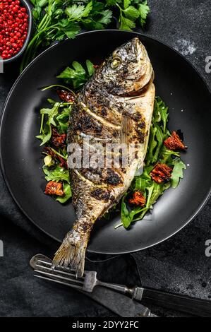 Gegrillter Dorado-Fisch mit Rucola-Salat und Tomaten. Schwarzer Hintergrund. Draufsicht Stockfoto