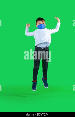 In voller Länge Foto von Vorschuljunge in einer medizinischen Maske, die auf einem grünen Hintergrund springt. Kid-Pandemiekonzept. Hochwertige Fotos Stockfoto