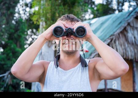 Vorderansicht eines zufriedenen jungen Mannes in einem weißen T-Shirt blickt durch ein Fernglas vor den Hintergrund einer Dorfhütte. Stockfoto