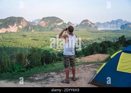 Ein junger Mann in einem weißen T-Shirt steht oben neben dem Zelt und blickt durch ein Fernglas in die Ferne, auf die Tierwelt. Stockfoto