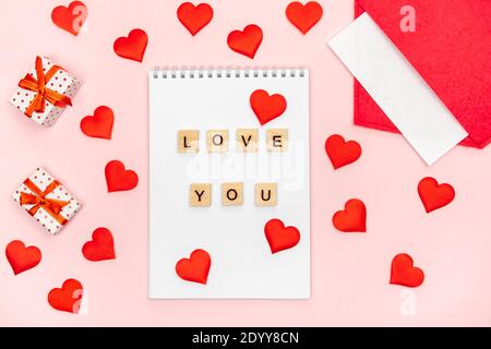 Flatlay Notizblock mit Holzbuchstaben I love you mit einem roten Umschlag mit Herzen und Geschenken auf rosa Hintergrund. Konzept des valentinstag und Schreiben Stockfoto