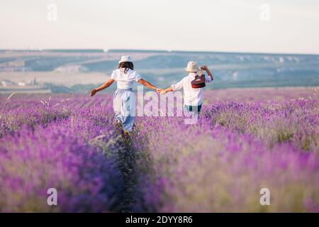 Junge und Mädchen von hinten in einem Lavendelfeld und rustikale Kleidung. Sie gehen mit einer Gitarre und halten die Hände fest Stockfoto