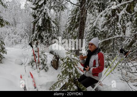 Frauen ruhen sich nach dem Skifahren im Wald aus, sitzen auf einem umgestürzten Baum mit einem verschneiten Wald im Hintergrund, Bild aus Vasternorrland Schweden. Stockfoto