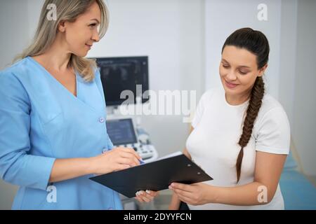 Geschickte Ärztin mit Klemmbrett in der Nähe von jungen schwangeren Frau während der medizinischen Diagnostik stehen. Lächelnd weiblich in Erwartung immer Untersuchung ar moderne Klinik. Stockfoto