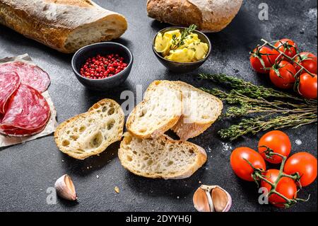 Zutaten für Sandwich mit Butter, geräuchertem Fleisch, Baguette, Kirschtomaten, Parmesankäse, Knoblauch und Gewürzen. Schwarzer Hintergrund. Draufsicht. Stockfoto