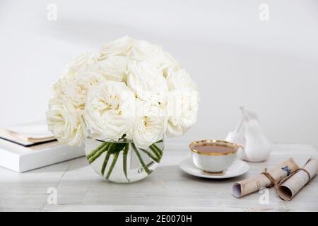 Ein Strauß weißer Rosen in einer Glasvase auf einem Tisch mit zwei Tassen Kaffee, einem Wasserkocher und einem Milchkännchen. Speicherplatz kopieren Stockfoto