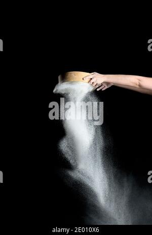 Weibliche Hand siebt weißes Weizenmehl durch ein rundes Holzsieb auf schwarzem Hintergrund, das Produkt wird gegossen Stockfoto