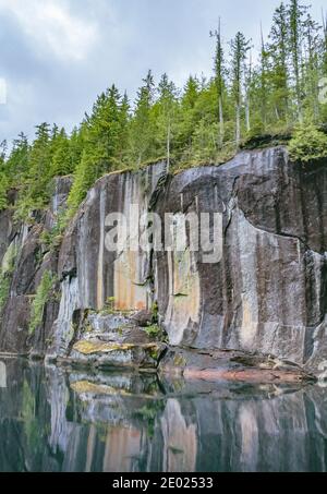 Gekrönt von dichtem Wald spiegeln sich steile senkrechte Klippen im stille Wasser darunter. Ein verblassenes Piktogramm ist sichtbar (Alison Sound, British Columbia). Stockfoto