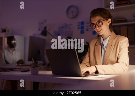 Junge Frau mit Brillen, die am Schreibtisch sitzt und an dem gearbeitet wird Geschäftsplan mit Laptop spät am Abend Stockfoto