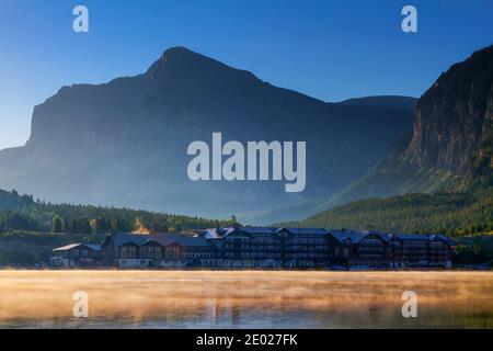 Das erste Licht des Sonnenaufgangs erwärmt die Landschaft rund um das Glacier Hotel, während der Nebel vom Swiftcurrent Lake zu den großen Frühaufstehern aufsteigt. Stockfoto