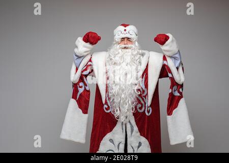 Weihnachtsmann mit erhobenen Händen in rotem Kostüm, ältere männliche grauhaarige Bärte auf grauem Studiohintergrund Stockfoto