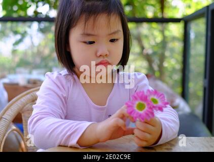 Ein nettes schüchternes asiatisches Mädchen, das an einem Tisch in einem Café sitzt, sich entspannt und ihre schönen rosa Blumen genießt, die verwirrt aussehen. Stockfoto